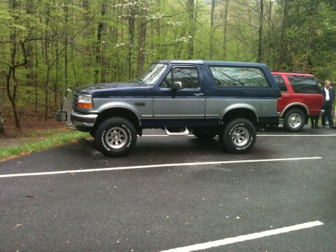 1992 Ford Bronco - This two-tone blue and silver 1992 Ford Bronco is parked alongside a red SUV in a wooded area.
