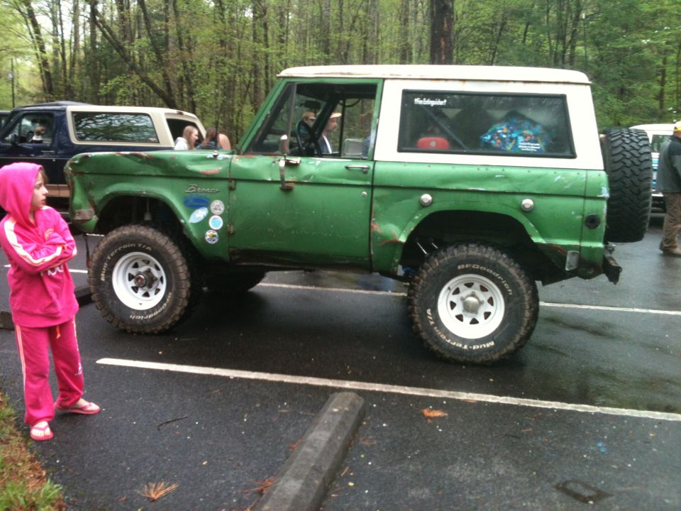 Green Ford Bronco with white top, parked next to a blue Bronco. A girl in a pink hoodie stands nearby