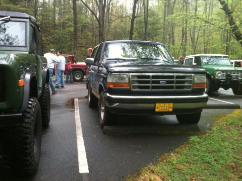 1992 Ford Bronco - A dark blue 1992 Ford Bronco is parked in a lot, with other vehicles and people visible in the background.