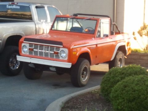 1987 Ford Bronco - This orange Ford Bronco is parked next to a silver pickup truck in front of a building.