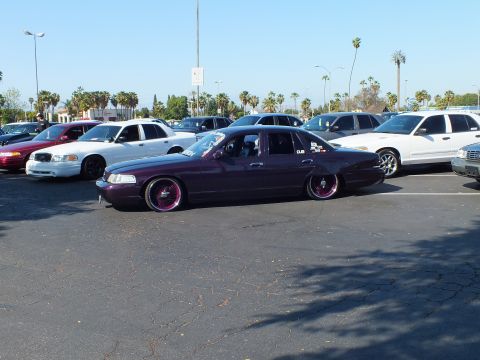 Slammed maroon 1990s sedan on pink-spoke wheels in a sunny parking lot