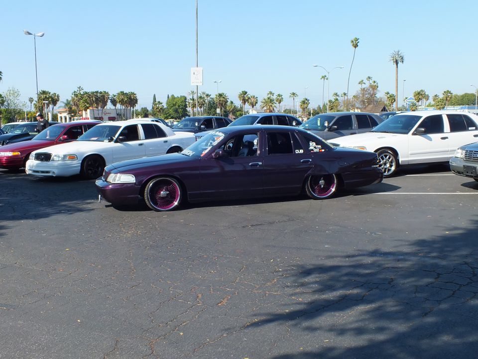 Slammed maroon 1990s sedan on pink-spoke wheels in a sunny parking lot
