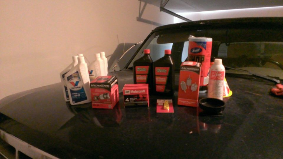 Bottles of oil, oil filters, spark plugs, and a roll of paper towels on a car hood