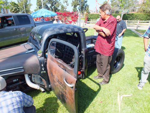 2003 Ford Crown Victoria - man in a red shirt stands inspecting a black rat rod truck with its rusted door open and hand-lettered signage.