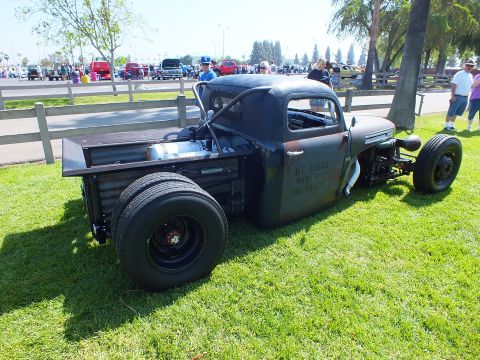 2003 Ford Crown Victoria - chopped black Ford rat rod truck with hand-painted lettering sits low on grass at a crowded outdoor car show.