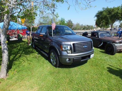 2003 Ford Crown Victoria - gray Ford F-150 crew-cab pickup flies a US flag on grass at a car show, with a black rat rod truck parked behind it.