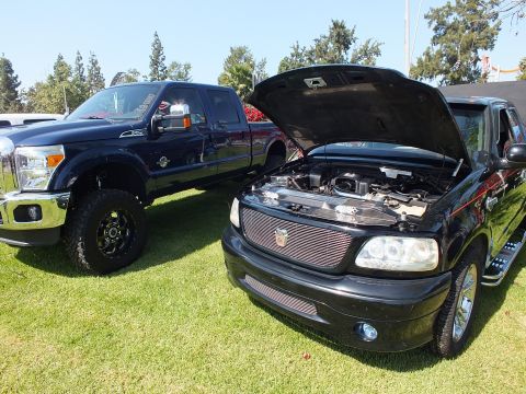 2003 Ford Crown Victoria - black Ford F-150 with raised hood and billet grille sits beside a lifted dark Ford Super Duty pickup on grass.