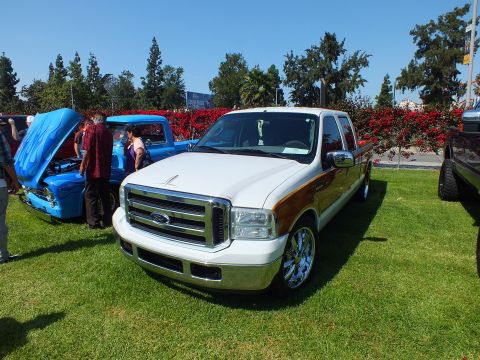 2003 Ford Crown Victoria - white Ford crew-cab pickup with wood-grain side panels and chrome wheels parked on grass at a car show, with a flamed blue truck nearby.