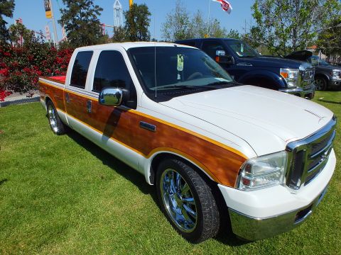 2003 Ford Crown Victoria - lowered white Ford crew-cab pickup with wood-grain side trim and chrome wheels parked on grass, other show trucks behind.