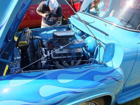 2003 Ford Crown Victoria - man leans over the engine bay of a blue flamed classic truck, photographing a polished chrome V8 under the open hood.