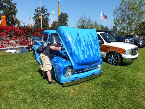 2003 Ford Crown Victoria - man bends into the open hood of a light-blue flamed classic Ford pickup at a sunny outdoor car show, amusement rides in the background.