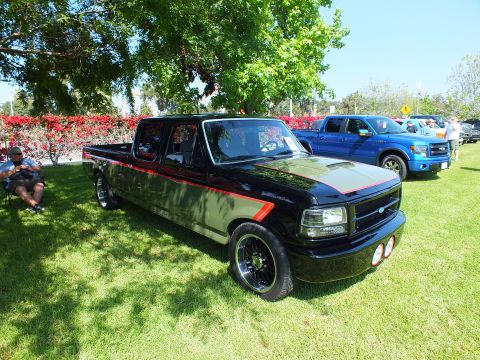 2003 Ford Crown Victoria - black lowered Ford crew-cab pickup with red striping on grass, a blue Ford F-150 and spectators visible behind it.