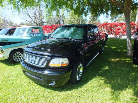 2003 Ford Crown Victoria - black Ford F-150 crew-cab pickup with a billet grille and chrome wheels parked on grass beside a teal classic truck at a show.