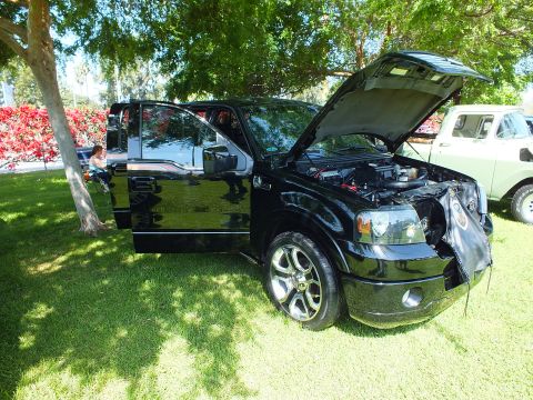 2003 Ford Crown Victoria - black Ford F-150 crew-cab pickup with open hood and driver door parked in tree shade on grass at an outdoor car show.