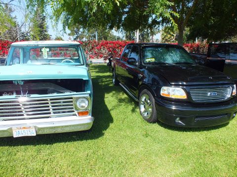 2003 Ford Crown Victoria - teal classic Ford pickup parked beside a black Ford F-150 with chrome billet grilles on a grassy car-show lawn.