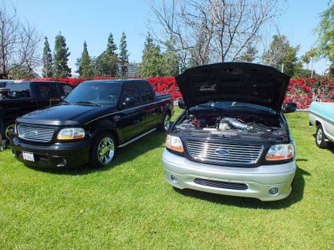 2003 Ford Crown Victoria - black Ford F-150 crew-cab parked beside a two-tone silver-and-black F-150 with an open hood on a grassy show field.