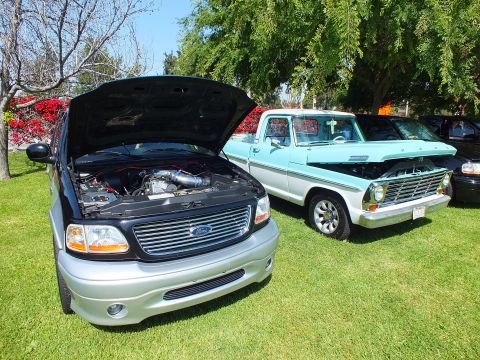 2003 Ford Crown Victoria - modern black Ford pickup displays its engine bay next to a restored light-blue classic Ford pickup on a grassy show field.