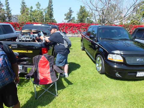 2003 Ford Crown Victoria - man works under the open hood of a black truck next to a lowered black Ford pickup with chrome wheels at an outdoor gathering.