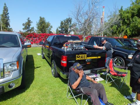 2003 Ford Crown Victoria - black custom pickup labeled MCE shows an exposed chromed engine while a seated man rests in a folding chair at the show.