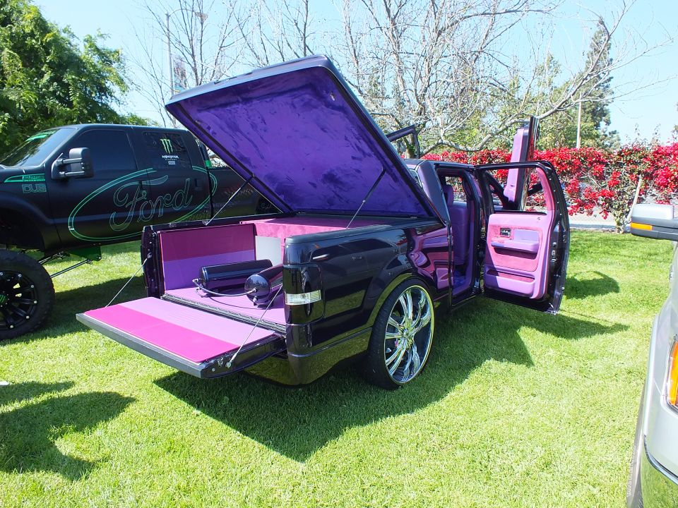 Custom black and pink-purple truck with raised hood-lid bed cover and open door on grass
