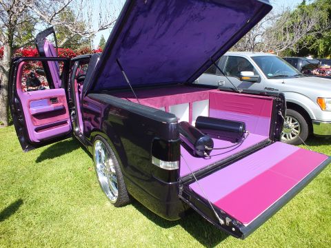 2003 Ford Crown Victoria - custom black truck displays its purple-lined raised lid and pink bed with doors open, parked beside a white Ford F-150.