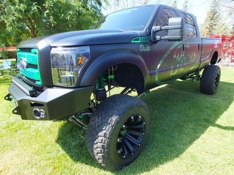 2003 Ford Crown Victoria - lifted matte-black Ford Super Duty with a teal grille, green suspension and black off-road wheels sits on a show lawn.