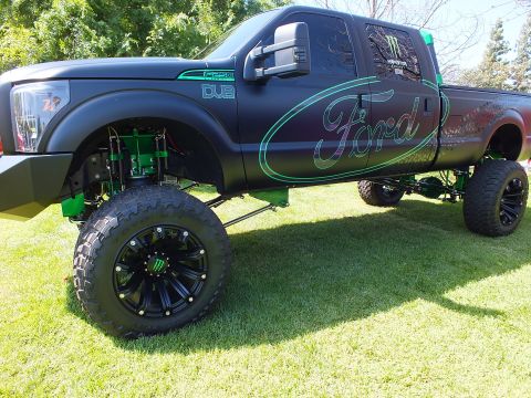 2003 Ford Crown Victoria - lifted matte-black Ford truck shows teal Ford script lettering, DUB badging and exposed green-painted suspension links.