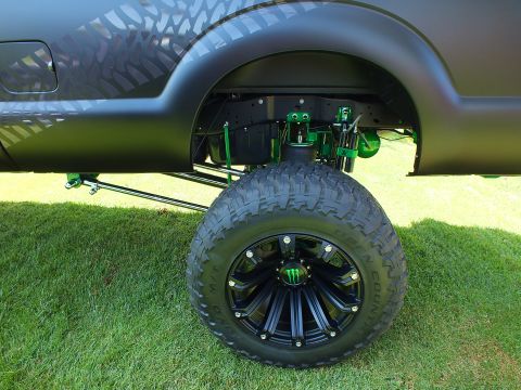 2003 Ford Crown Victoria - lifted matte-black truck's front axle is exposed, showing green suspension links and a black off-road wheel under the fender.