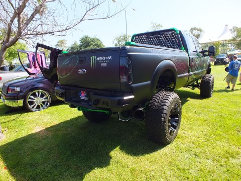 2003 Ford Crown Victoria - matte-black lifted Ford truck with Monster Energy badging sits at a show, a purple custom truck visible to its left.