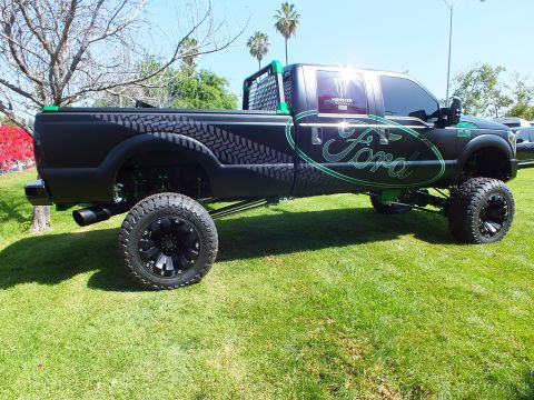 2003 Ford Crown Victoria - long lifted matte-black Ford truck shows teal Ford script and tire-tread side graphics, raised high on black wheels.