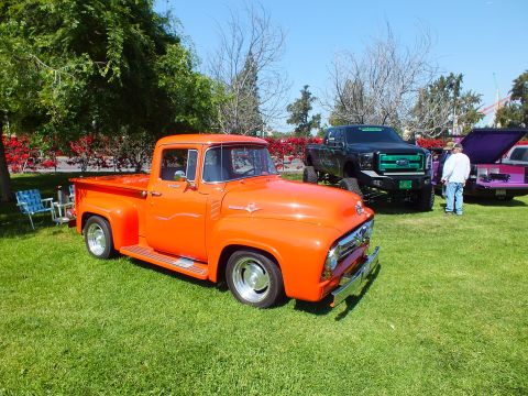 2003 Ford Crown Victoria - orange restored vintage Ford pickup parked on grass next to a lifted black truck and a purple vehicle at a car show.