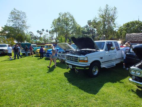 2003 Ford Crown Victoria - white lifted Ford pickup with its hood up displayed among other classic trucks and onlookers at a grassy car show.