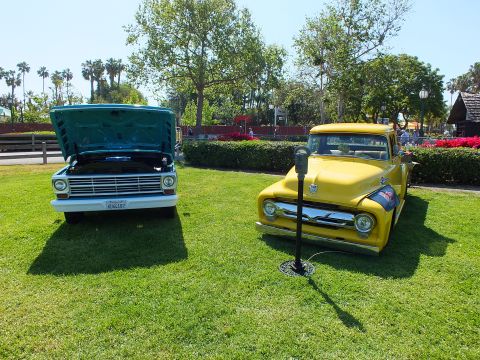 2003 Ford Crown Victoria - teal pickup with raised hood sits next to a glossy yellow vintage Ford pickup on a green lawn at an outdoor car show.