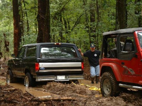 1995 Ford Bronco - Two off-road vehicles, a 1995 Ford Bronco and a red Jeep, are stuck in a muddy trail in the woods.