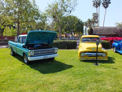 2003 Ford Crown Victoria - teal Ford pickup with its hood up parked beside a yellow vintage pickup on a lawn with red flowers and palm trees behind.