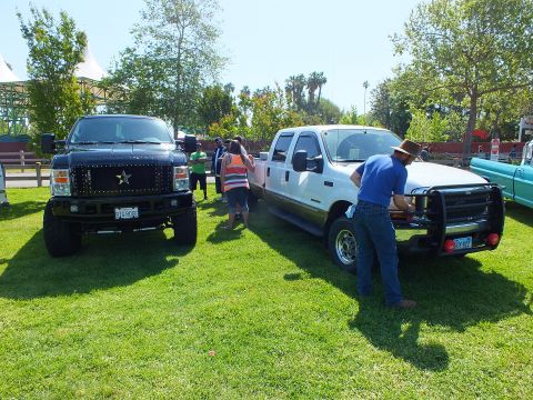 2003 Ford Crown Victoria - black lifted truck with a star grille and a white crew-cab pickup parked on grass while attendees inspect them at a show.