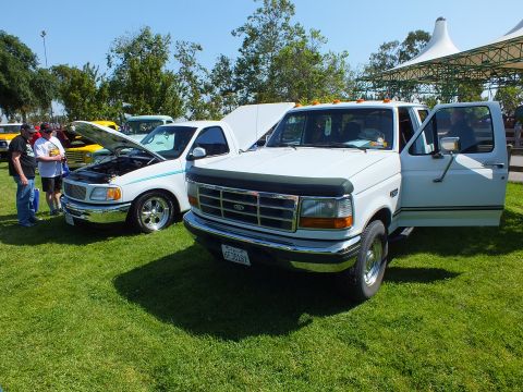 2003 Ford Crown Victoria - white Ford crew-cab pickup with bug shield and open door parked beside a lowered truck under a canopy at a car show.