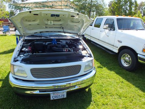2003 Ford Crown Victoria - white truck with raised hood, billet grille and CRUSZR plate sits beside a white Ford crew-cab pickup on a show lawn.