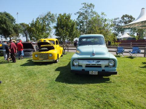 2003 Ford Crown Victoria - light blue vintage Ford pickup beside a yellow vintage Ford pickup on a lawn with attendees and a canopy in the background.