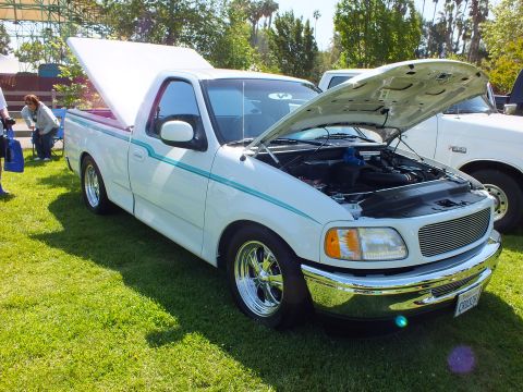 2003 Ford Crown Victoria - white lowered Ford pickup with raised hood, billet grille and teal pinstriping displayed on a lawn at an outdoor car show.