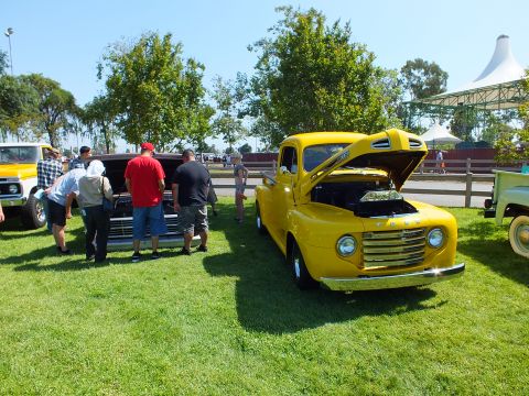 2003 Ford Crown Victoria - bright yellow restored vintage Ford pickup with raised hood on grass while a group of people examine a nearby truck.