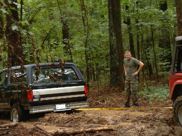 Ford Bronco stuck in mud with a man standing nearby and part of a red Jeep visible