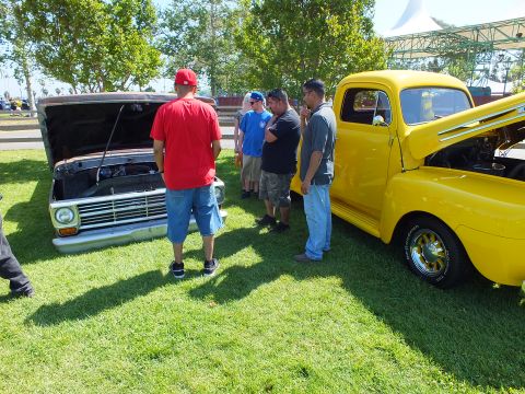 2003 Ford Crown Victoria - group of men stand between a dark hood-up pickup and a glossy yellow vintage Ford pickup on a lawn at a car show.