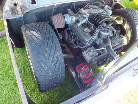 2003 Ford Crown Victoria - Close view into a vintage pickup engine bay showing a supercharged V8 with a red cone air filter, red battery box and a wide front tire.