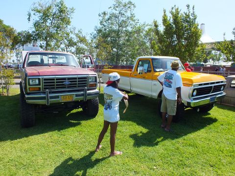 2003 Ford Crown Victoria - maroon squarebody Ford 4x4 and a yellow 1970s Ford pickup sit on show grass while spectators look at the trucks.