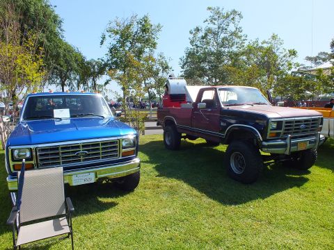 2003 Ford Crown Victoria - blue 1980s squarebody Ford pickup parked beside a lifted maroon Ford 4x4 on sunny show grass with trees behind.