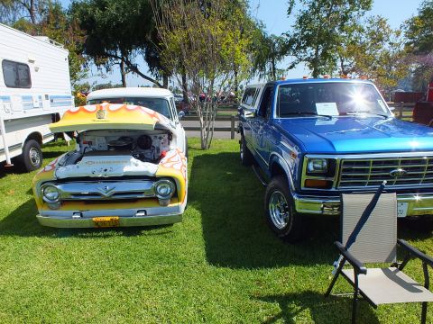 2003 Ford Crown Victoria - white-and-yellow flame-painted 1950s Ford F-100 with its hood up sits beside a blue squarebody Ford pickup at a car show.