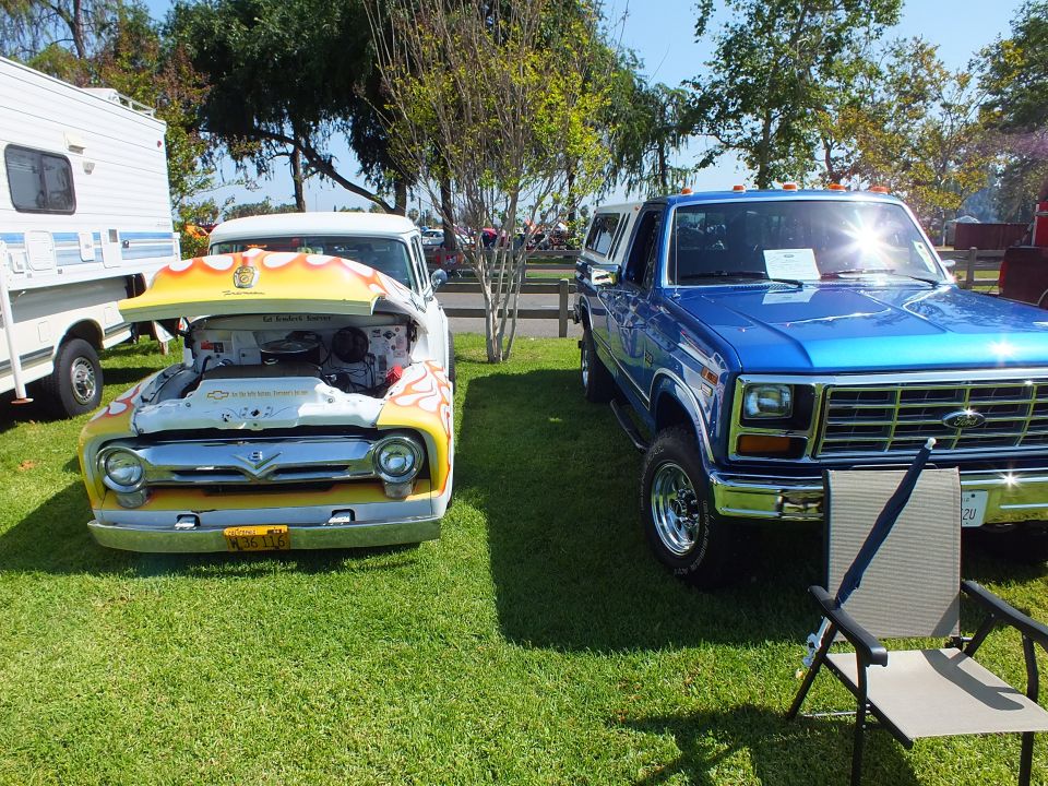 flame-painted 1950s Ford pickup with open hood beside a blue 1980s Ford pickup on grass