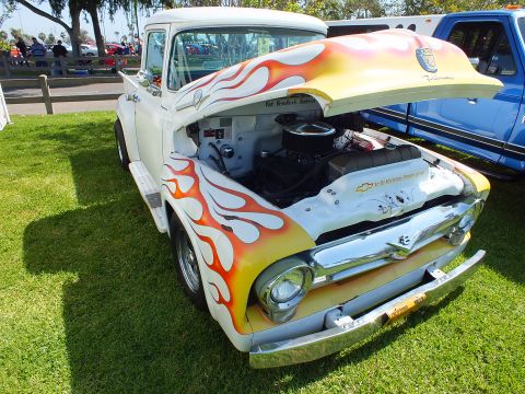2003 Ford Crown Victoria - white 1950s Ford F-100 with orange-yellow flames sits hood-up on show grass, exposing a chromed V8, beside a blue pickup.