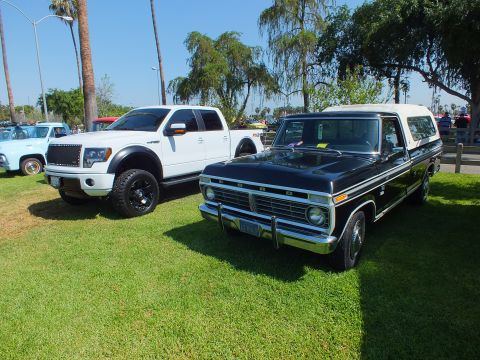 2003 Ford Crown Victoria - white modern lifted Ford F-150 sits beside a black 1970s Ford pickup with a white canopy at a sunny car show.